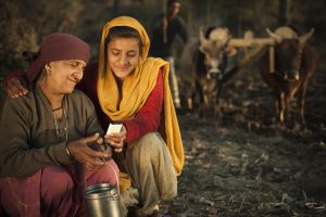 Real people from rural India: Happy peasant girl showing mobile phone to her grandmother and her elder brother plowing agricultural field in the background with the help of oxes. The image is shot in the real location of a village of Himachal Pradesh, India.